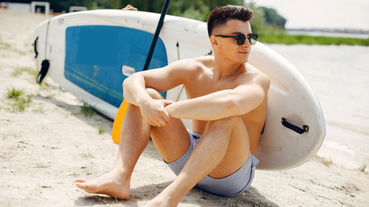 Tide And Style -Tide And Style What to Wear to the Beach Man leaning on surfboard with sunglasses and boardshorts